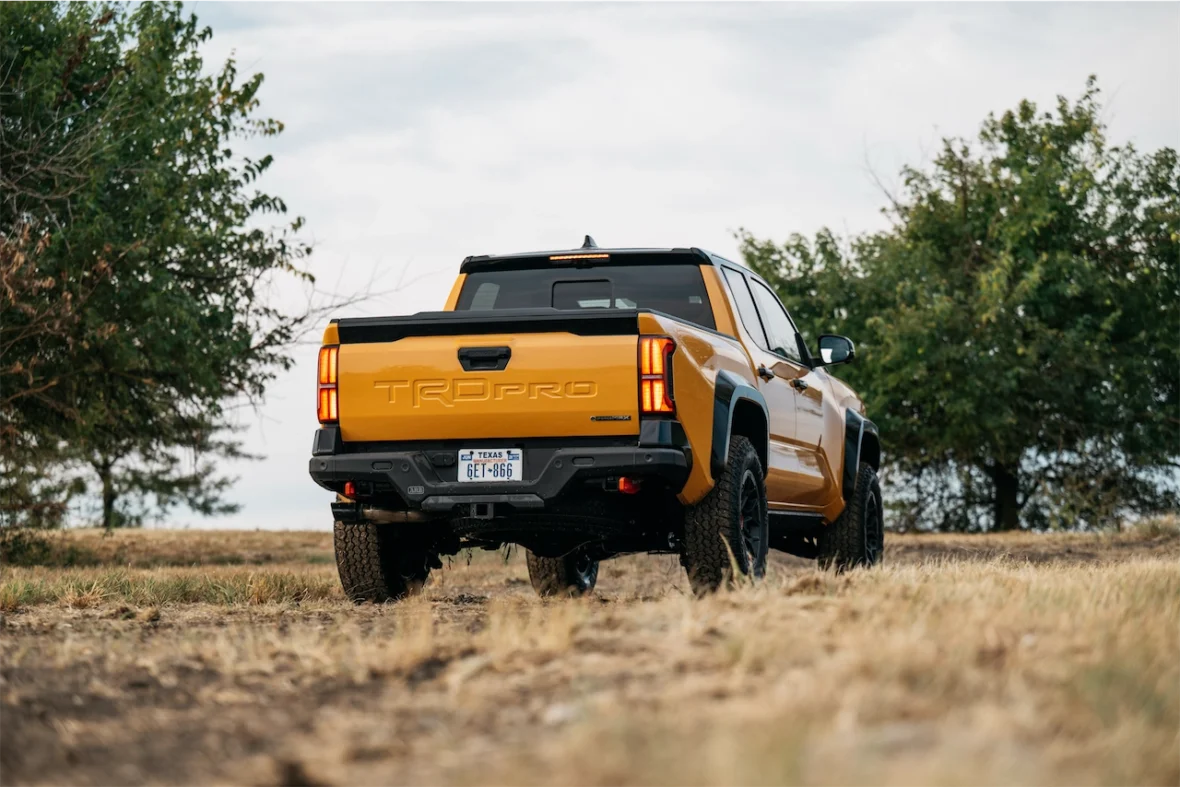 Rear view of a yellow TRD Pro truck parked on a dirt path, surrounded by trees and grass under a cloudy sky.