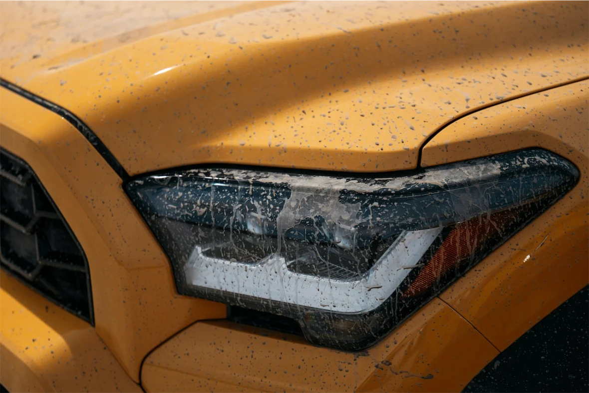 Close-up of a yellow vehicle's headlight and hood covered in dirt and water spots.