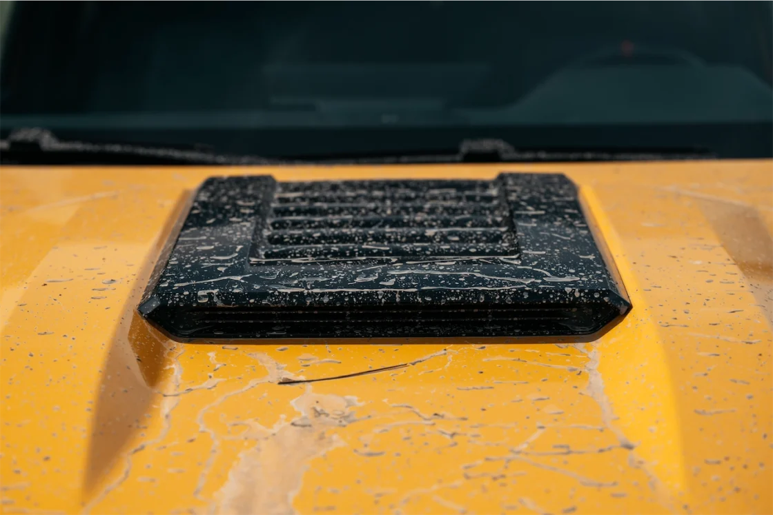 Close-up of a dirty yellow vehicle hood with a black vent cover, showing splashes of mud and water droplets.